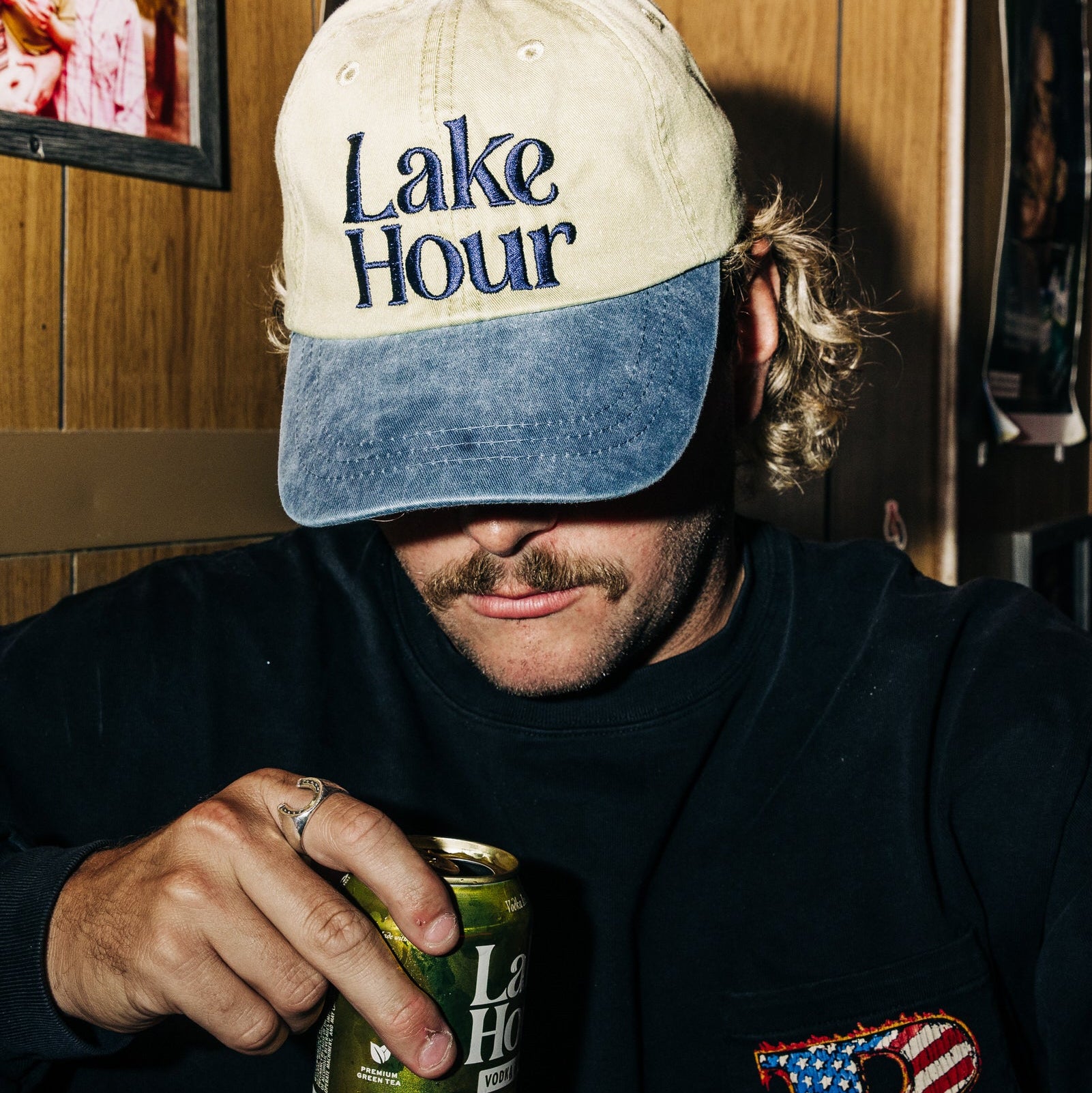 Man wearing a 'Lake Hour' cap and holding a can, with a wooden wall background
